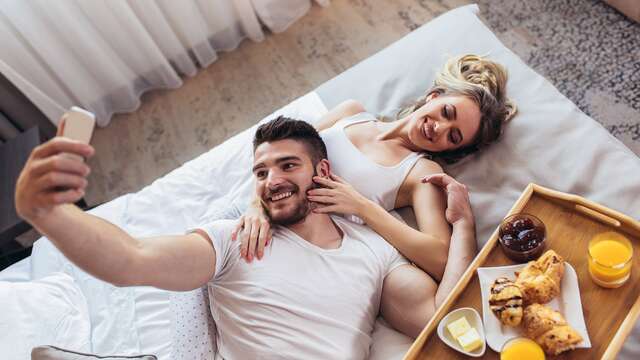"Couple prenant un selfie au lit avec un plateau petit-déjeuner, un moment parfait d'un citytrip en Europe. "