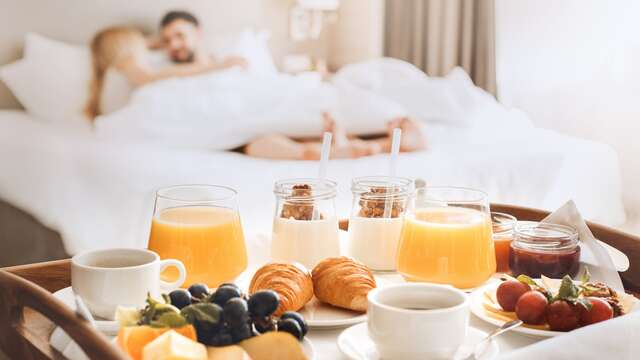 Bandeja de desayuno a los pies de una cama donde descansa una pareja, en una luminosa habitación de hotel.