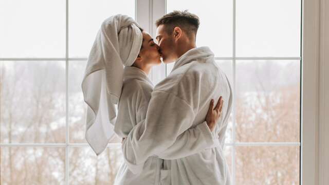 Pareja en albornoz abrazándose en su habitación mientras miran por la ventana un paisaje nevado.