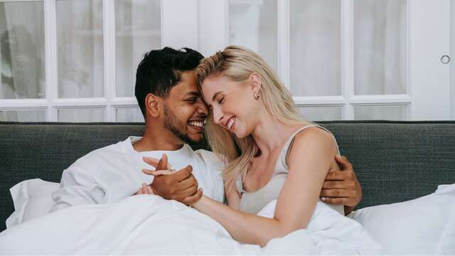 Pareja sonriente y abrazada sobre la cama de una espaciosa y luminosa habitación de hotel de lujo.
