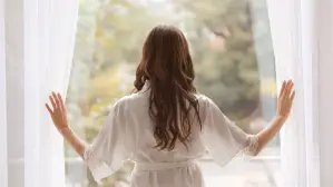 Woman in white robe opens curtains to a bright green outdoor view.