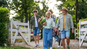 A family of four walks through a white garden gate, carrying flowers.