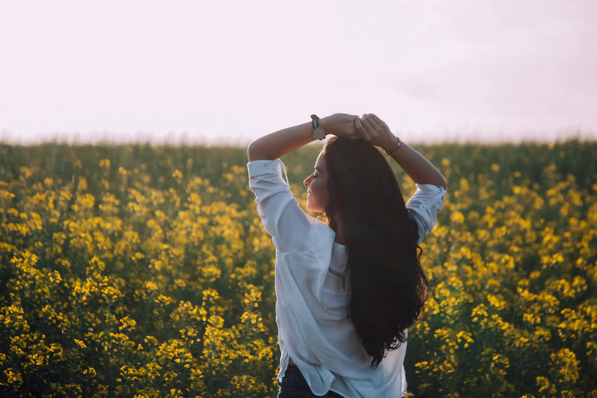 Woman with long dark hair standing in a field of yellow flowers at sunset, arms raised.