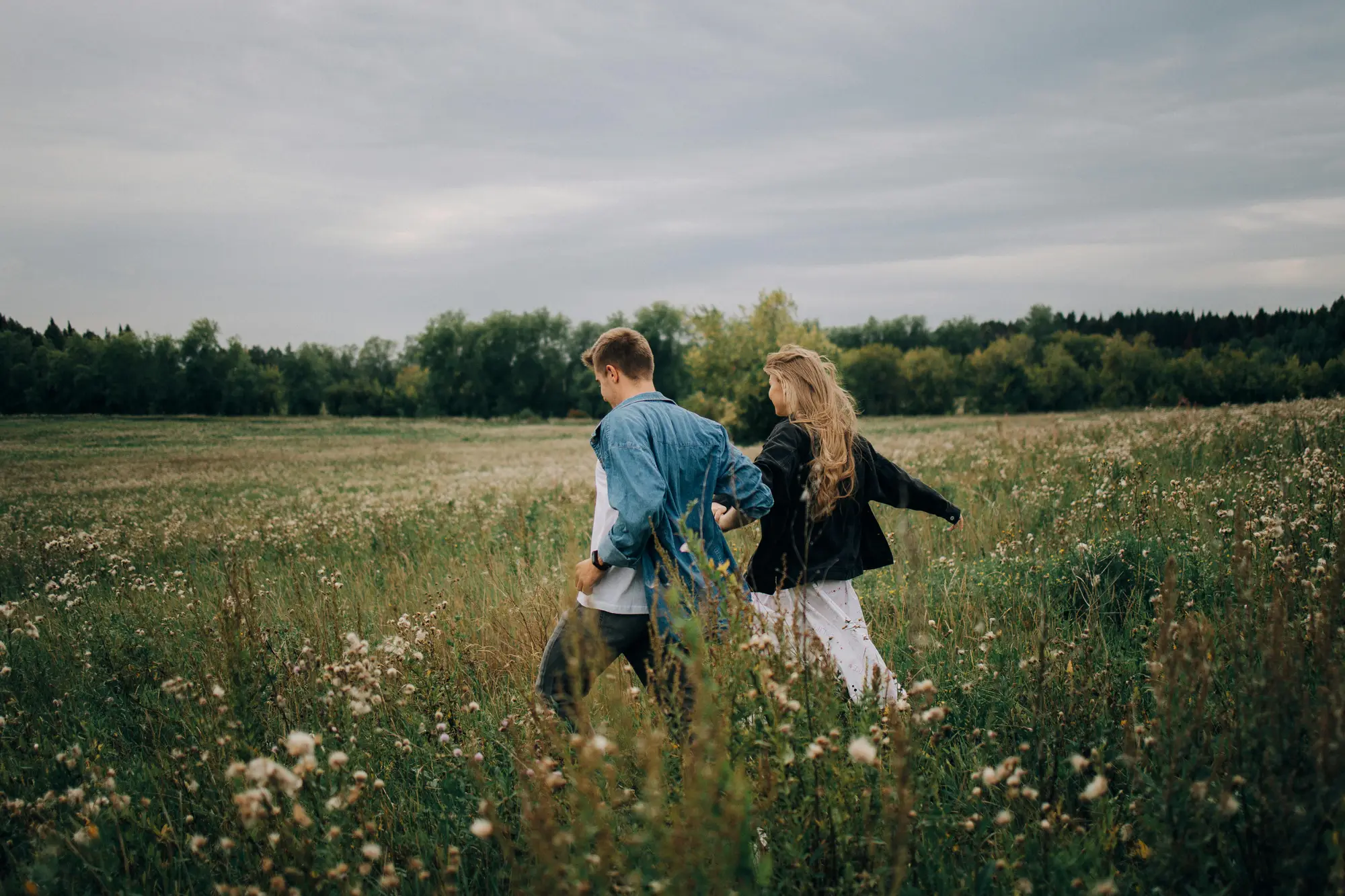 A young couple walks hand-in-hand through a field of wildflowers under a cloudy sky.