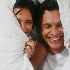 Happy couple playfully peeking from under a soft white blanket, smiling brightly.