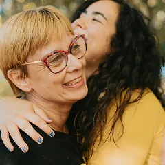 Two happy women, one with blonde hair and glasses, embrace with warm smiles.