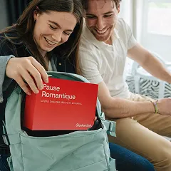 Young woman puts red 'Pause Romantique' box into backpack, smiling man looks on.