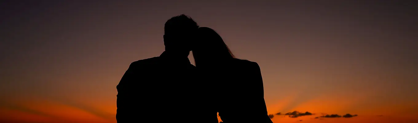 Silhouettes of a couple with their heads together against a vibrant sunset sky.