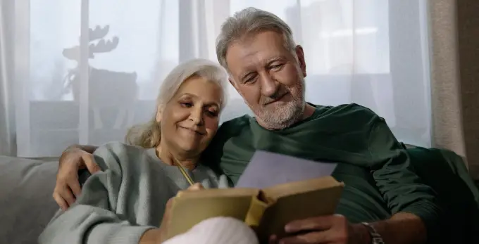 Elderly couple smiling, reminiscing over old photos in an album on their couch.