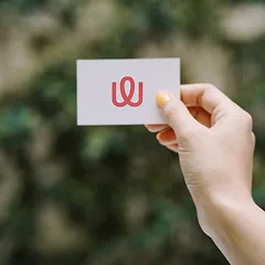 Hand holding a white business card with a red 'W' logo against blurred greenery.
