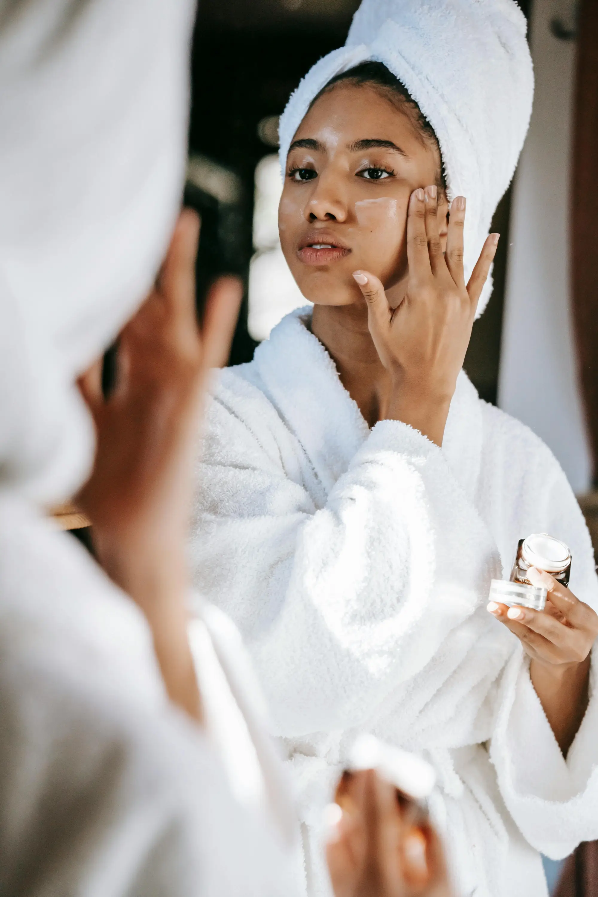 Woman in white bathrobe applying face cream while looking at her reflection in a mirror.